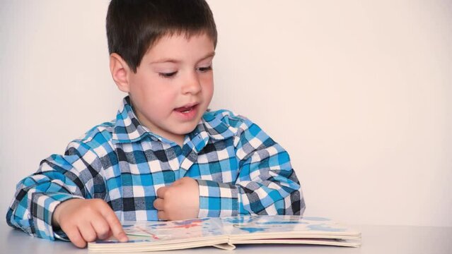 A 4-year-old boy looks at a book with thick cardboard pages with pictures, talks on a white background.