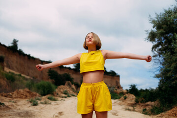 A young girl with her eyes closed, in a yellow summer suit, shorts and a tank top, stands among the...