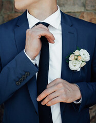 A stylish groom in a blue suit and black tie, in a white shirt, straightens his tie and looks to the side. Morning preparations on the wedding day. - obrazy, fototapety, plakaty