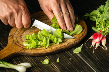 Close-up of a chef hands cutting green lettuce leaves on a cutting board with a knife for preparing a vegetarian dish. Peasant foods.