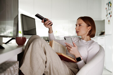 Woman watching tv, using remote control and phone, channel surfing, getting information and latest news, watching movies, bored at home. Reading book. Weekend and free time, social media and network