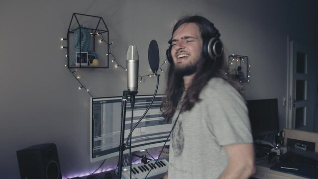 Man with long hair pretending to play air guitar in headphones with a microphone and audio recording software in the background at his home music studio.