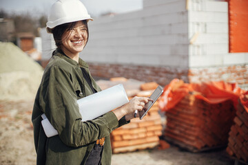 Happy young female architect with tablet checking blueprints against of building new modern house....