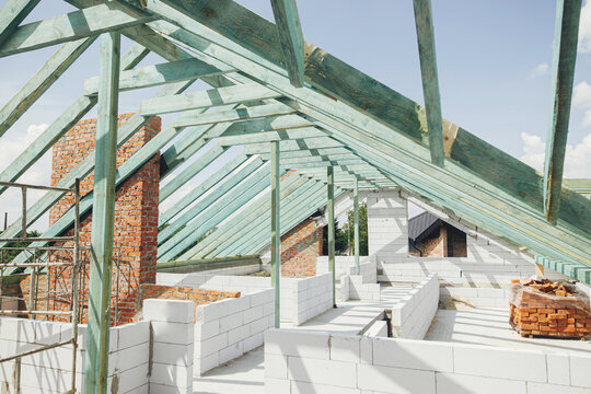 Unfinished roof trusses, view in attic with rafters, beams, windows and chimney. Wooden roof framing. Mansard of modern farmhouse building construction.