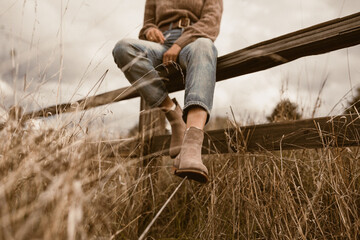 Close-up shot of boots of a woman sitting on a wooden fence in a rustic field - shoe advertisement