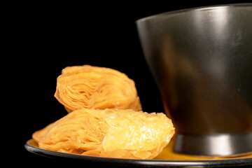 Two pieces of sweet honey baklava on a saucer with a black cup, close-up, isolated on a black background.