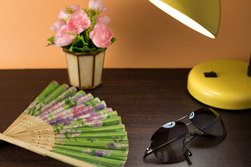 detail concept of bedside table with luxury sunglasses, fan, flowers and a lighted lamp