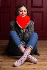 Smiling short-haired woman in jacket sitting on floor legs crossed holding heart shape in front of her chin