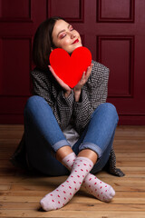 Dreamy smiling short-haired woman in jacket sitting on floor legs crossed holding heart shape in front of her chin