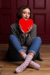 Short-haired woman in jacket looking into camera sitting on floor legs crossed holding heart shape in front of her chin