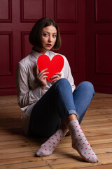 Short-haired woman looking into camera sitting on floor legs crossed holding heart shape in front of her