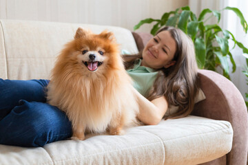 happy beautiful girl playing with dog at home. pomeranian playing with owner. love to animals. human pet friendship, adoption. young cute woman hugging spitz, lying on sofa