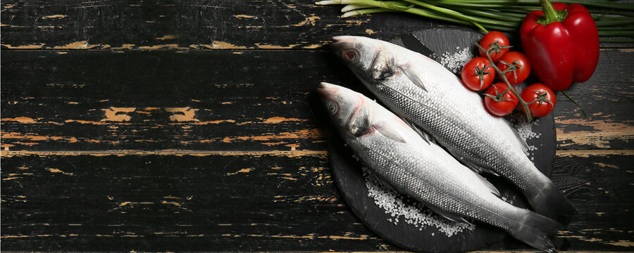Fresh uncooked sea bass fish and vegetables on black wooden background with space for text
