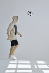 Young sportsman in street style wear training with soccer ball in large bright white hall with...