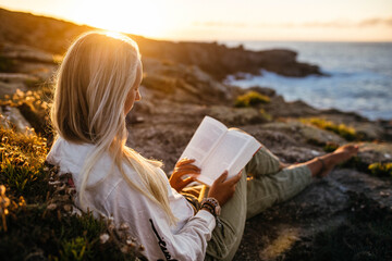 Hispanic female sitting at the rocky beach reading a book at sunset