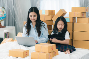 Young busy business woman and mother sitting with daughter on bed surrounded by parcel cardboard box while using laptop and digital tablet