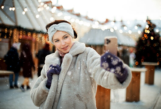 Charming young woman in winter clothes at the Christmas market takes a selfie on a smartphone
