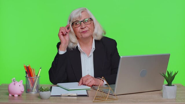 Thoughtful senior businesswoman making notes, writing down thoughts with pen into notepad notebook diary, to do list, good idea sits at workplace at office. Elderly woman on green chroma key studio
