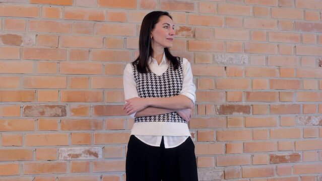 Elegant thoughtful caucasian businesswoman in houndstooth knit vest and white shirt stand with crossed arms. Woman posing over brick wall background. Close-up