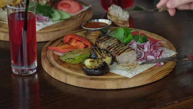 A woman in a restaurant with a knife and fork eats the national dish lula-kebab with vegetables and baked potato slices with pita bread on a wooden platter. Close-up, no face
