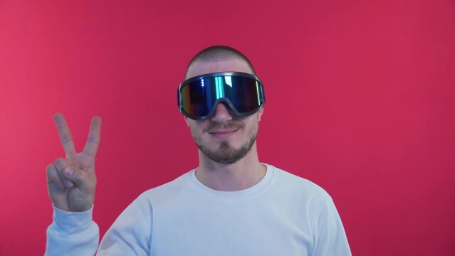 Snowboarder Shows Two Figers, Peace Sign. Man in Snowboard Goggles in a White Jacket on a Red Background in the Studio