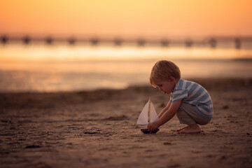 Little boy playing on the sunset sea beach with toy ship. Concept of childhood and family vacation. adventure and travel