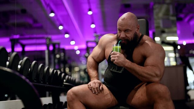 A mature male bodybuilder drinks an energy drink made of fruit during a break in the gym. A male athlete drinks celery energy drink after physical education in the gym.