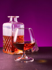 glass textured square bottle and glass goblet with amber alcohol brandy scotch whiskey bourbon. violet background