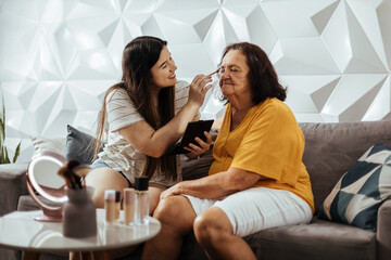 Neta adulta aplicando maquiagem em sua avó. Avó e neta se divertindo em casa.