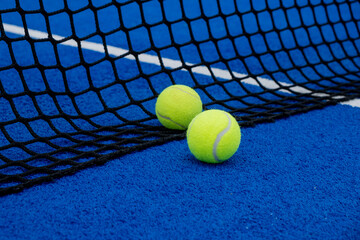 Two balls near the net of a blue artificial grass paddle tennis court.