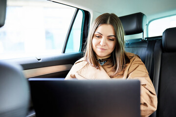 Attractive business woman working on her laptop at the back sit of a car on the way to meeting. Side view of beautiful and confident woman in coat working on laptop while sitting in the car.
