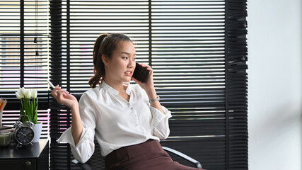 Millennial young business woman sitting at comfortable workplace and talking on mobile phone