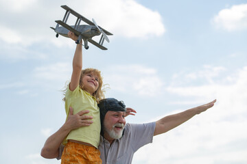 Young grandson and old grandfather having fun with plane outdoor on sky background with copy space. Child dreams of flying, happy childhood with grand dad.