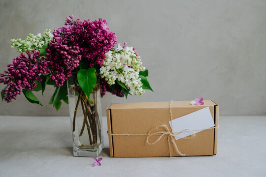 Craft box tied with twine on a bow. A gift and a branch of lilac. Delivery of goods to the door. The package is on a gray background.