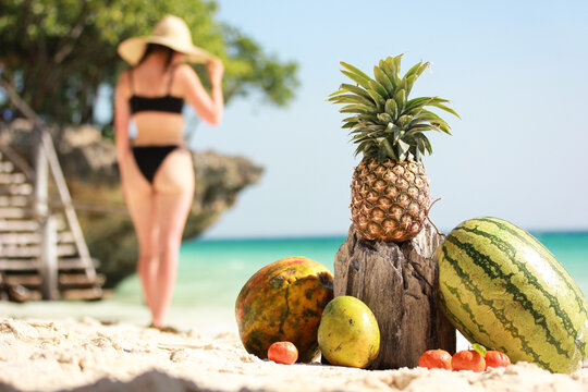A beautiful girl standing at a carribean island beach with tropical fruits laying on the white sand and ocean  coconut pineapple mango watermelon and papaya on a hot summer day blue sky holiday season