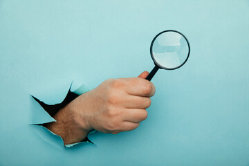 Male hand holds black magnifying glass in torn hole of blue background. Searching information...