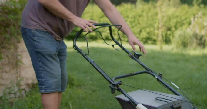 A man in the garden repeatedly pulls sharply on a cable to start a power mower. Medium shot