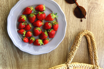 Lilac plate full of fresh strawberries, straw bag and retro sunglasses on wooden table. Flat lay.