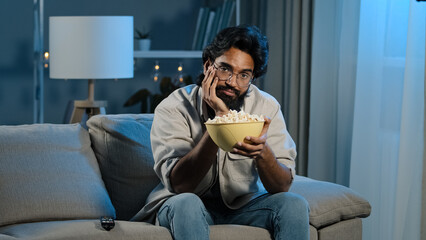 Upset sad bored Arab man tired indian male with beard sits at home evening watching boring TV program switches channel laughing enjoying comedy movie eating popcorn relaxing in dark living room alone