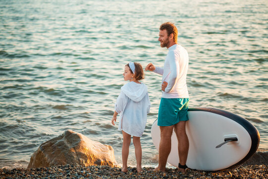 Man and preschool girl standing on the beach with sup board. Ocean's surface on the background. Summer active sport at the sea. Back view