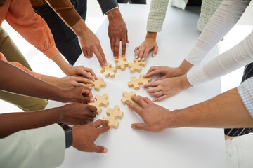 Diverse group of young mixed race multiethnic people joining pieces of wooden jigsaw puzzle on office desk as metaphor for business team and teamwork. Cropped shot of human hands holding jigsaw parts