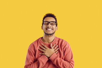 Happy black man in glasses smiling and holding his hands on his chest to express endless gratitude and appreciation. Cheerful ethnic guy says thank you from the bottom of his heart for gift or present - obrazy, fototapety, plakaty