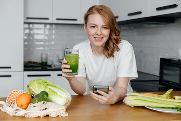 Young caucasian woman drinking fresh green smoothie uses smartphone to count calories in kitchen