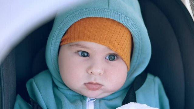 Calm peaceful beautiful baby boy wearing warm clothes sits in a pram. Close up portrait of a toddler at walk.