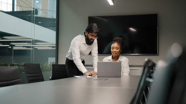 Professional Arabian Indian teacher mentor helping African woman new employee teaching intern explaining online project service corporate app using laptop. Teamwork internet win discussion in office