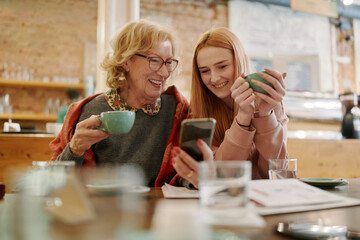 Happy grandmother and her adolescent granddaughter sitting in a cafe, enjoying coffee and using smart phone for funny videos. - 505654237