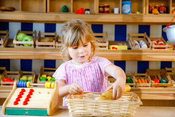 Little preschool girl play with food and grocery wooden toys. Happy active child playing role game as cashier or seller, in wood shop or supermarket. Education, activity for kids.