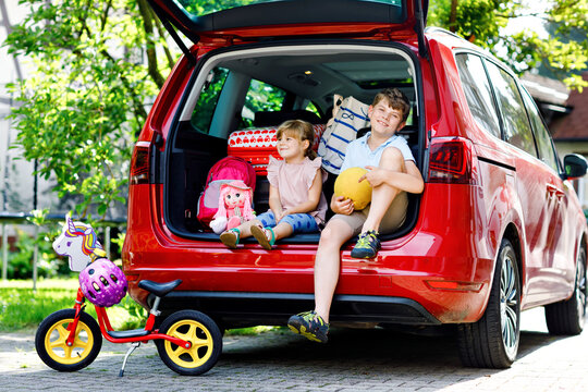 Two children, school boy and preschool girl sitting in car trunk before leaving for summer vacation with parents. Happy kids, siblings, brother and sister with suitcases and toys going on journey