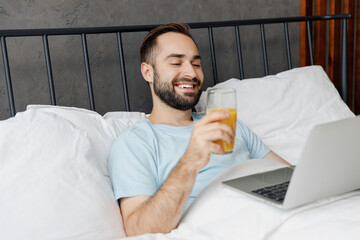 Young smiling man in blue t-shirt lying in bed hold use work on laptop pc computer drink juice est...