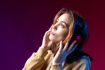 Young beautiful fashionable hipster girl dressed in a hoodie  listening to music in red headphones in a studio red dramatic background